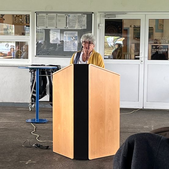 Eine ältere Frau steht an einem Rednerpult und spricht zu einem Publikum. Hinter ihr spiegeln Glastüren einen Raum mit einem Dartboard und Personen wider. Ein Tisch mit Taschen steht in der Nähe.