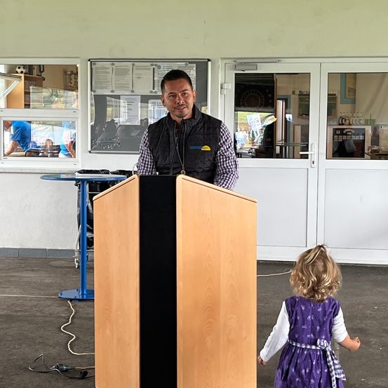 Ein Mann steht hinter einem Rednerpult mit einem Mikrofon und spricht eine Zuhörerschaft an. Hinter ihm geht ein kleines Mädchen auf ihn zu. Im Hintergrund befinden sich ein Tisch und Türen.