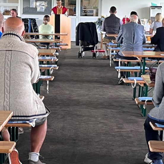 Eine Gruppe von Menschen sitzt an Tischen in einem Saal. Eine Person steht an einem Podium. Tische und Stühle sind in Reihen angeordnet. Einige Personen tragen Jacken. Ein Kinderwagen steht in der Nähe des Eingangs.