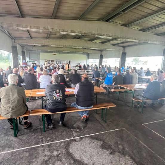 Eine große Gruppe von Menschen sitzt auf Bänken in einem großen, offenen Raum. Einige haben Papiere, und eine Rednertribüne ist in der Mitte aufgestellt. Der Raum hat große Fenster und einen Betonboden.