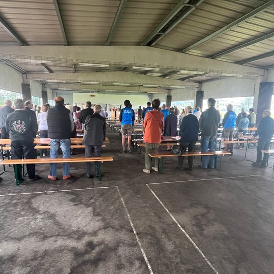 Ein großer Innenbereich mit mehreren Holzbänken, wo Menschen stehen und sitzen, in unterschiedlicher Kleidung gekleidet. Der Bereich ist gut beleuchtet mit Lampen an der Decke.