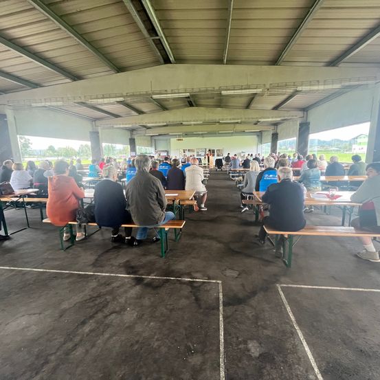 Eine Gruppe von Menschen sitzt auf Bänken in einem großen Saal. Einige tragen Turnschuhe, während andere Sandalen tragen. Einige Tische haben Papiere darauf. Der Saal hat ein großes Glasfenster an der Seite.