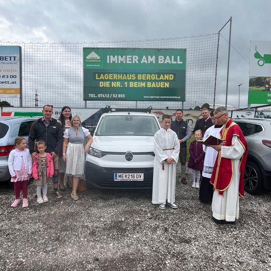 Ein Priester segnet eine Familie vor einem weißen Auto mit dem Kennzeichen ME 216 DV. Dahinter befindet sich ein Banner, das für Lagerhaus Bergland wirbt.