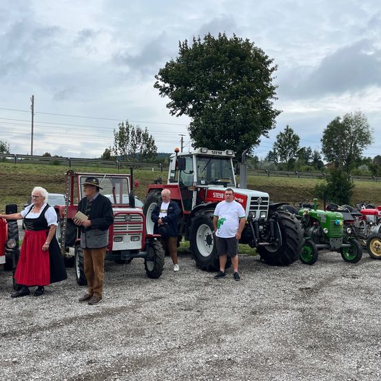 Eine Gruppe von Menschen steht vor alten Traktoren, einer hält ein Buch. Die Traktoren sind auf Kies geparkt, mit einem Baum und einem bewölkten Himmel im Hintergrund.