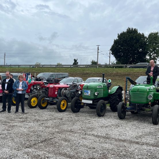 Eine Gruppe von Menschen steht neben alten Traktoren auf einem Kiesplatz unter einem bewölkten Himmel.