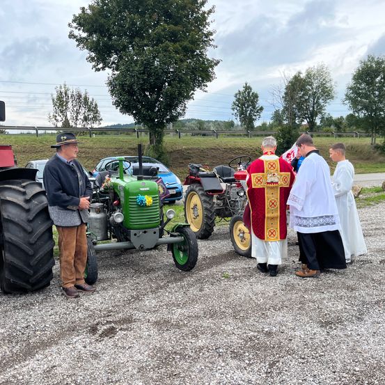 Ein Mann steht neben einem grünen Traktor, während zwei Priester sprechen. Hinter ihnen ist ein weiterer Traktor geparkt, mit Autos und einem Zaun im Hintergrund.
