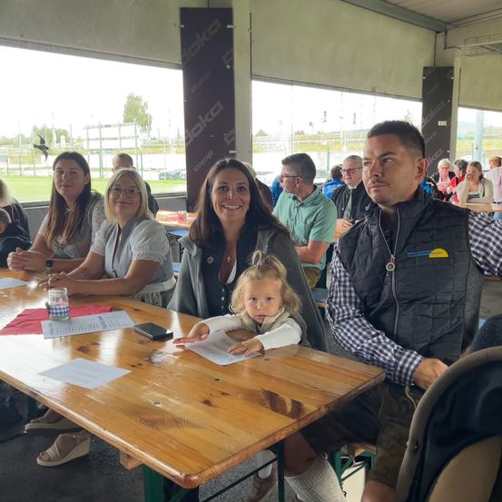 Eine Gruppe von Menschen sitzt in einem Pavillon an einem Tisch und lächelt für ein Foto. Eine Frau hält ein Baby, und ein Mann sitzt neben ihnen. Papiere und ein Handy liegen auf dem Tisch. Glasfenster bieten einen Blick auf ein grasiges Feld.