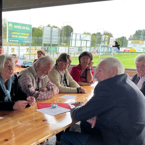 Eine Gruppe älterer Personen sitzt an einem Tisch und führt ein Gespräch. Sie befinden sich in einem Außenbereich mit einem großen Fenster hinter ihnen, das ein Rasenfeld zeigt.