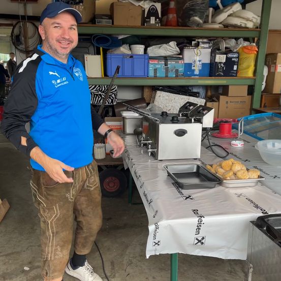 Ein Mann in einem blauen Shirt und braunen Hosen steht in einem überfüllten Raum, lächelt und gestikuliert mit seinen Händen. Hinter ihm steht ein Tisch, der mit einem weißen Tuch bedeckt ist und ein silbernes Gerät und eine Schüssel mit Essen aufweist. Regale und Kisten sind im Hintergrund zu sehen.