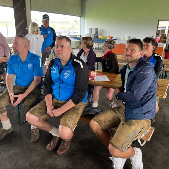 Fünf Männer in traditioneller Kleidung sitzen auf Bänken in einem Raum. Ein Mann trägt einen Hut. Hinter ihnen unterhalten sich mehrere Personen. Ein Tisch hat eine Kerze und Papiere. Glasfenster zeigen eine Aussicht nach draußen.