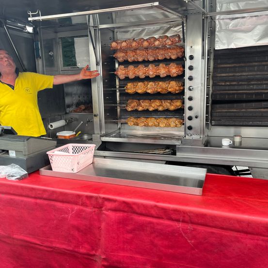 Ein Mann in einem gelben Shirt bedient einen großen gewerblichen Ofen mit Hähnchenspießen im Inneren. Ein rosa Korb und ein Edelstahltablett liegen auf dem Tresen.
