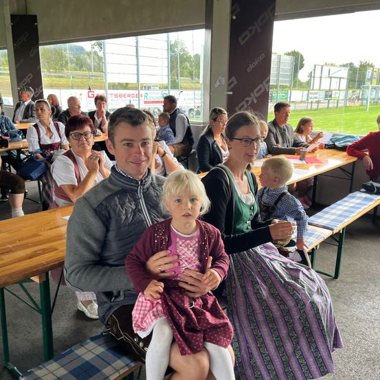 Eine vierköpfige Familie sitzt an einem Tisch in einem Restaurant. Der Mann hält ein kleines Mädchen, während eine Frau neben ihm ein traditionelles Kleid trägt. Sie sitzen in der Nähe eines großen Fensters mit Blick auf ein Feld und ein Auto in der Ferne.