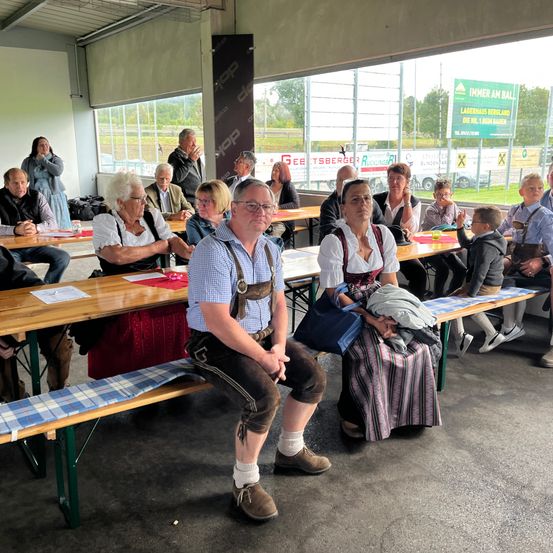Eine Gruppe von Menschen, möglicherweise auf einem Festival, sitzt auf Bänken in einem Raum mit Fenstern. Sie tragen traditionelle Kleidung. Ein Mann sitzt vorne, während andere hinter ihm sitzen. Papiere liegen auf den Tischen.
