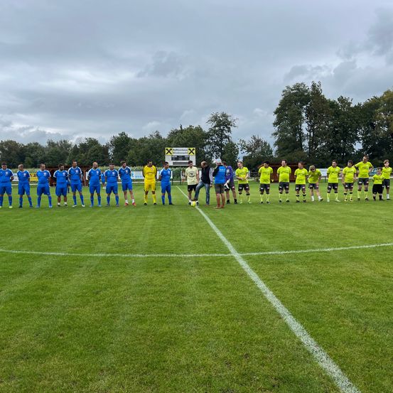 Zwei Fußballteams stehen auf einem Feld, sich gegenüberstehend. Eine Mannschaft trägt Blau, die andere Gelb. Sie sind von einer weißen Linie und Bäumen umgeben.