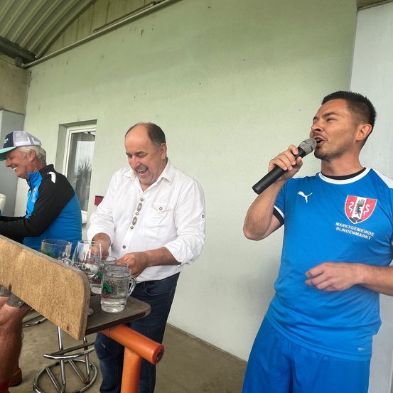 Ein Mann in einem blauen Trikot mit einem Logo auf der rechten Seite singt in ein Mikrofon. Ein weiterer Mann steht hinter ihm mit einem Lächeln im Gesicht. Sie befinden sich in einem Raum mit einer weißen Wand und einem Tisch mit Gläsern vor ihnen.