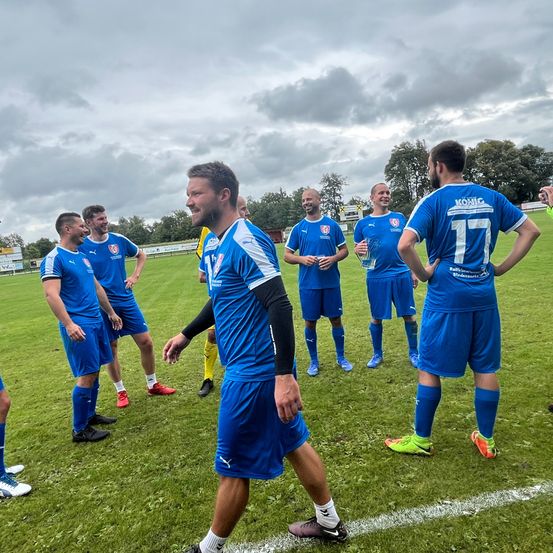 Eine Gruppe männlicher Fußballspieler in blauen Trikots steht auf einem Feld. Ein Spieler hält eine Trophäe. Sie alle schauen nach vorne und scheinen zu reden.