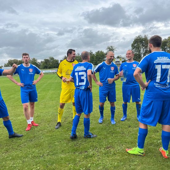 Fußballspieler in blauen Trikots, einer mit der Nummer 13, unterhalten sich auf einem Rasenfeld. Einer hält ein Glas, während andere Getränke in der Hand haben. Bewölkter Himmel und Bäume im Hintergrund.