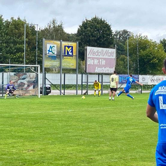 Ein Fußballspiel ist im Gange. Spieler in blauen und gelben Trikots sind auf dem Feld. Ein Spieler tritt den Ball, während andere in Position sind.