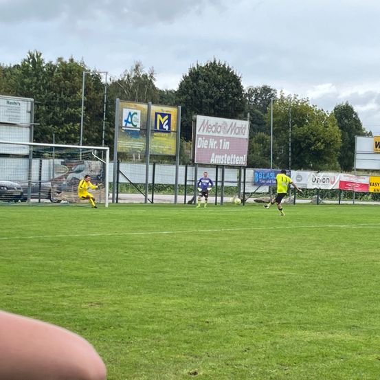 Ein Fußballspiel findet auf einem Spielfeld statt, bei dem zwei Teams spielen. Die Spieler befinden sich in verschiedenen Positionen. Im Hintergrund befinden sich mehrere Werbetafeln und ein Zaun.
