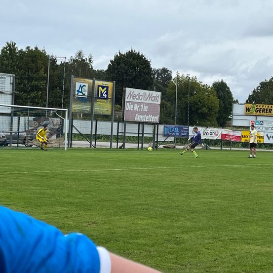 Ein Fußballspiel findet auf einem Feld mit Werbung an der Umzäunung statt. Die Spieler tragen gelbe Trikots, und ein Spieler ist kurz davor, den Ball zu schießen.