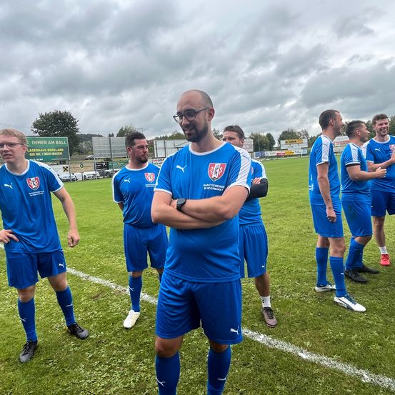 Eine Fußballmannschaft in blauen Trikots steht auf einem Rasenfeld. Der Mann vorne hat die Arme verschränkt. Sie bereiten sich wahrscheinlich auf ein Spiel vor.