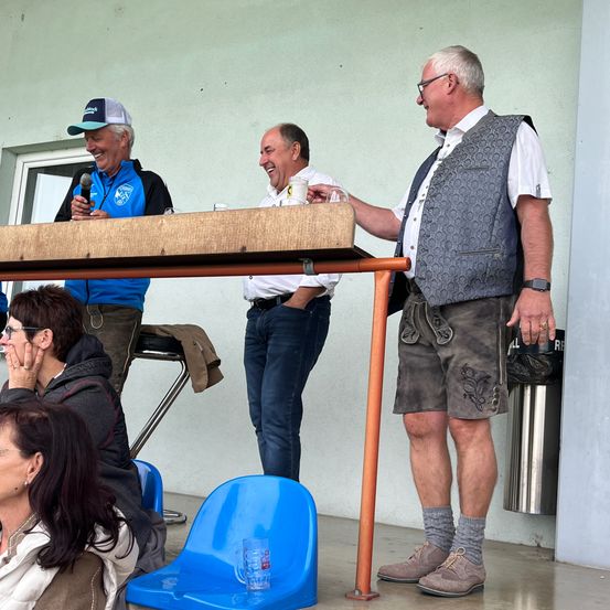 Drei Männer in Freizeitkleidung stehen bei einem langen Tisch. Einer hält ein Glas, ein anderer ein Mikrofon und der dritte hat eine Jacke auf dem Schoß. Hinter ihnen sitzen Menschen auf Stühlen. Ein Mülleimer steht neben der Wand.