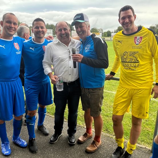 Fünf Männer, vier in Fußballtrikots, stehen auf einem Feld. Der Mann in der Mitte hält ein Mikrofon und scheint zu sprechen. Die Männer links und rechts lächeln.