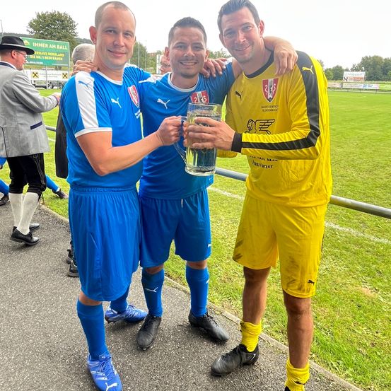 Drei Männer in Sporttrikots stehen auf einem Weg, einer hält einen großen Glasbecher, wahrscheinlich zur Feier eines Sieges. Hinter ihnen ist ein Rasenfeld mit einem Zaun und ein Schild links.