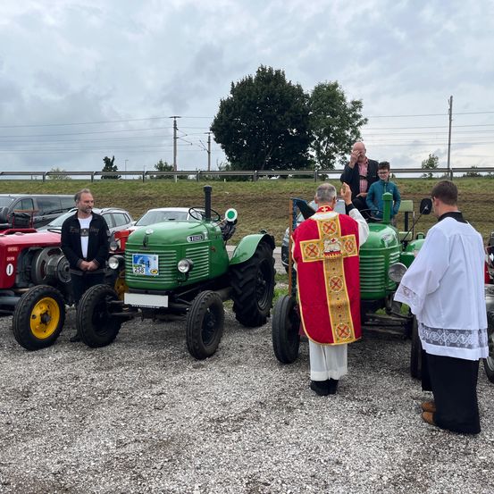 Ein Priester segnet vor einer Reihe alter Traktoren auf einer Kiesstraße. Ein Mann sitzt auf einem Traktor, während andere in der Nähe stehen.