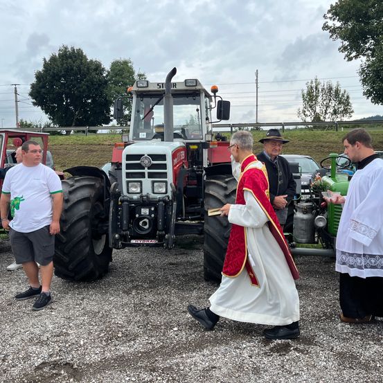Zwei Priester stehen neben einem Traktor mit einem Mann in einem weißen T-Shirt. Dahinter steht ein Mann mit einem schwarzen Hut und einer Brille.