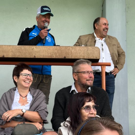Eine Gruppe von Menschen versammelt sich in einem Stadion. Ein Mann steht, hält ein Mikrofon und lächelt. Ein weiterer Mann in einem braunen Jackett lächelt ebenfalls. Zwei Frauen und ein Mann sitzen davor und tragen eine Brille.