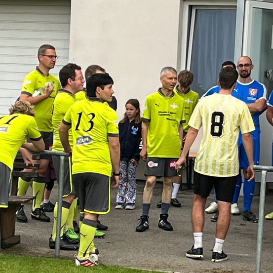 Eine Gruppe von Männern in gelben Trikots steht vor einem Gebäude, ein Mann in einem gelb-weißen Trikot mit der Nummer 8 steht ihnen gegenüber, in der Nähe eines Mädchens in einer blauen Jacke.