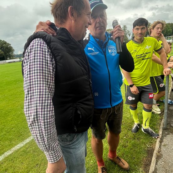 Ein älterer Mann spricht in ein Mikrofon, ein Mann in einem blauen Jackett steht neben ihm, beide auf einem Rasenfeld, mit jungen Spielern in grünen Trikots dahinter.