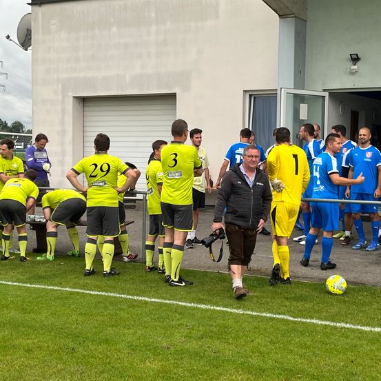 Eine Fußballmannschaft in gelben Trikots versammelt sich auf dem Feld, möglicherweise in Vorbereitung auf ein Spiel. Ein Mann in einer schwarzen Jacke geht mit einer Kamera. Das Feld hat eine weiße Linie.