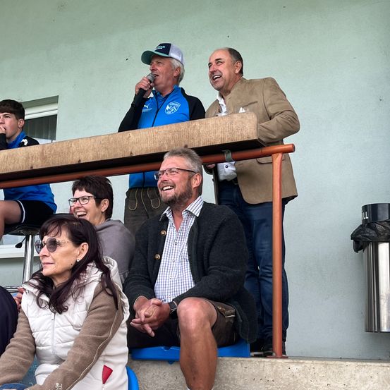 Eine Gruppe von Menschen sitzt in den Bänken, zwei Männer stehen hinter ihnen. Ein Mann hält ein Mikrofon und scheint zu sprechen. Der andere Mann lächelt. Hinter ihnen steht rechts eine Mülltonne.