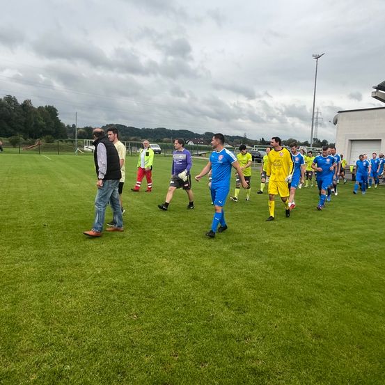 Eine Gruppe von Fußballspielern in blauen und gelben Trikots läuft auf einem Feld, möglicherweise auf dem Weg zum Spielfeld. Es stehen zwei Personen am Rand des Feldes.