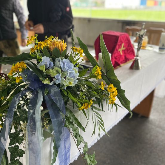 Eine farbenfrohe Blumenarrangement aus blauen und gelben Blumen steht auf einem mit einem weißen Tuch bedeckten Tisch. Ein rotes Tuch mit einem Kreuz befindet sich hinter den Blumen. Zwei Personen stehen im Hintergrund.