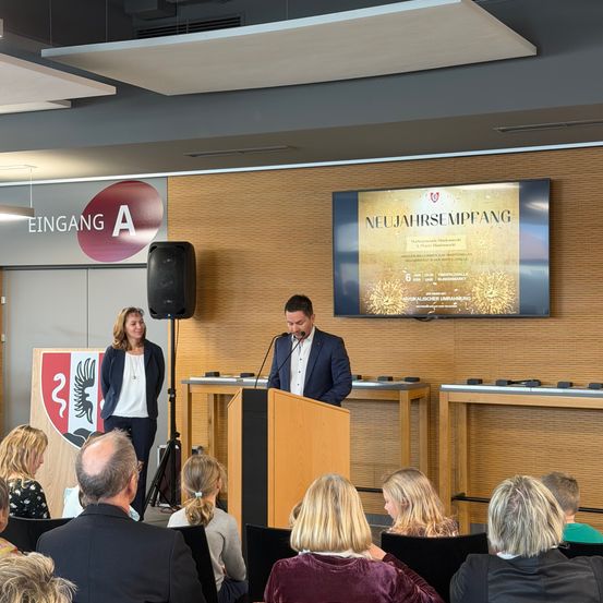 Ein Mann spricht an einem Podium vor einem Schild, das 'Neujahrsempfang' sagt. Eine Frau steht neben einem Lautsprecher. Leute sitzen in Stühlen vor ihnen.