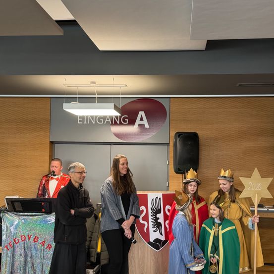 Mehrere Personen versammeln sich in einem Raum, wahrscheinlich für eine Veranstaltung. Zwei Frauen halten ein Sternenschild mit der Nummer 2026, während ein Mann in ein Mikrofon spricht. Eine Frau steht neben einem Banner mit einem Wappen. Ein Schreibtisch mit Laptop und Lautsprecher steht vor ihnen.