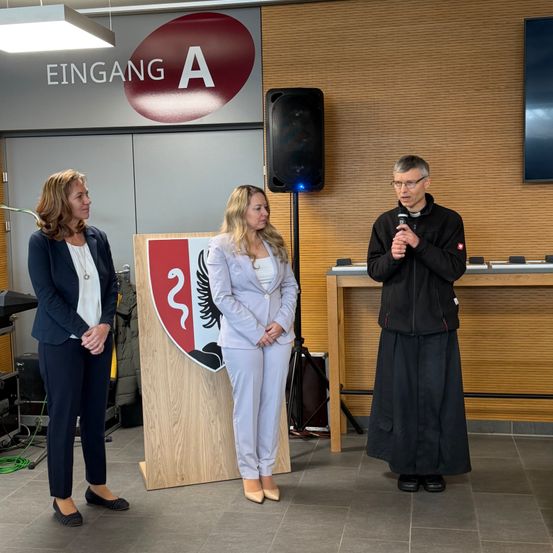 Drei Personen stehen neben einem hölzernen Podest in einem Raum. Ein Mann spricht in ein Mikrofon. Ein Schild mit der Aufschrift 'EINGANG A' ist darüber angebracht.
