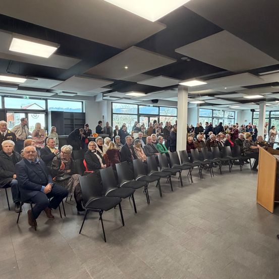 Eine große Versammlung von Menschen in formeller Kleidung sitzt in Reihen, vor einem Podium mit Mikrofon. Der Raum ist modern eingerichtet und hat große Fenster.