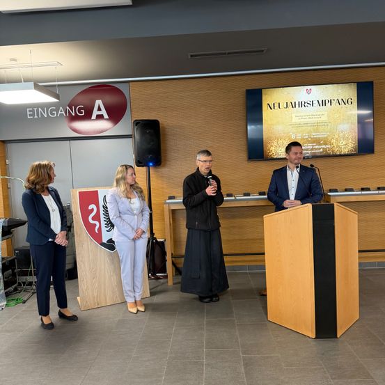 Vier Personen stehen in einem Konferenzraum. Ein Mann spricht an einem Podium mit Mikrofon. Hinter ihm zeigt eine Projektionsleinwand 'NEUJAHRSEEMPFANG'. Links steht ein Schild mit 'EINGANG A'.