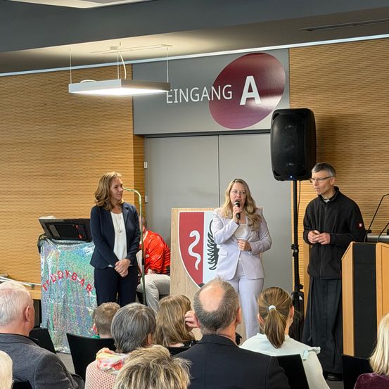 Eine Frau spricht in ein Mikrofon an einem Podium, während eine andere Frau hinter ihr steht. Ein Mann steht neben einem Lautsprecher auf der rechten Seite. Sie stehen vor einem Schild, das 'Eingang A' sagt. Ein Publikum sitzt auf Stühlen.