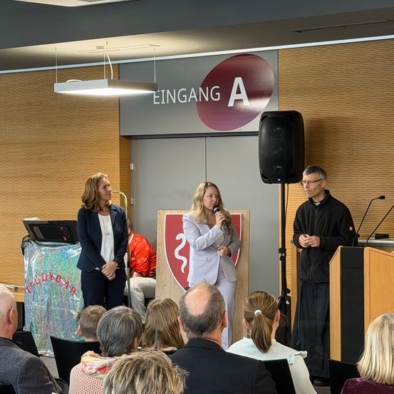 Drei Personen stehen vor einer Menschenmenge, wobei eine Frau in ein Mikrofon spricht. Dahinter befindet sich ein rot-weißes Schild. Ein erwachsener Mann steht neben einem Podium mit einem Mikrofon auf einem Ständer.