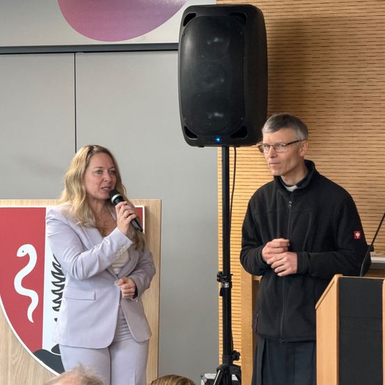 Eine Frau spricht in ein Mikrofon, während sie neben einem Mann vor einer hölzernen Wand mit einem Lautsprecher steht.