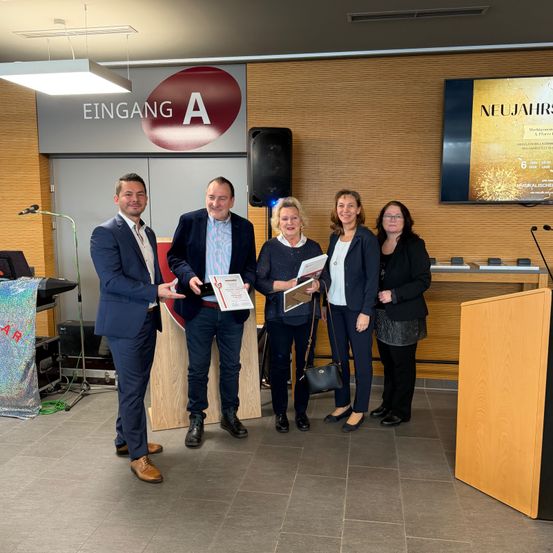 Fünf Personen stehen in einem Raum mit einem Podium, Mikrofonen und einem Banner mit der Aufschrift 'EINGANG A'. Sie sind in formeller Kleidung gekleidet, halten Urkunden in den Händen und lächeln. Hinter ihnen befindet sich eine Wand mit einer Projektionsleinwand und einem Lautsprecher.