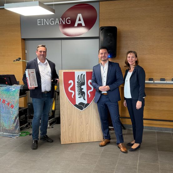 Drei Personen stehen vor einem Podium in einem modernen Raum. Der Mann auf der linken Seite hält ein Buch und ein Zertifikat, und der Mann in der Mitte hält seine Hände zusammen. Die Frau auf der rechten Seite trägt eine Halskette. Hinter ihnen steht ein Schild mit der Aufschrift 'Eingang A'.
