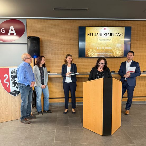 Eine Gruppe von Menschen steht vor einem Podium und einem Schreibtisch mit einer großen Leinwand dahinter, auf der 'Neujahrsempfang' steht. Der Mann am Podium hält ein Papier in der Hand.