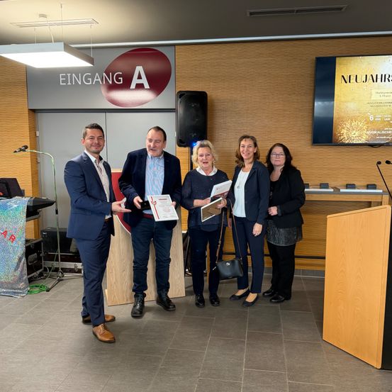 Fünf Personen stehen lächelnd in einem Raum. Sie halten Urkunden in den Händen und scheinen bei einer Preisverleihung zu sein. Ein Podium und ein großer Bildschirm sind hinter ihnen sichtbar.