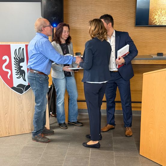 Eine Gruppe von Menschen steht in einem Raum, vielleicht einem Konferenzraum. Einer von ihnen hält einen Teller mit Essen. Der Mann vorne schüttelt einer Frau die Hand. Ein anderer Mann hält ein Buch. Der Raum hat Holzwände und -böden.